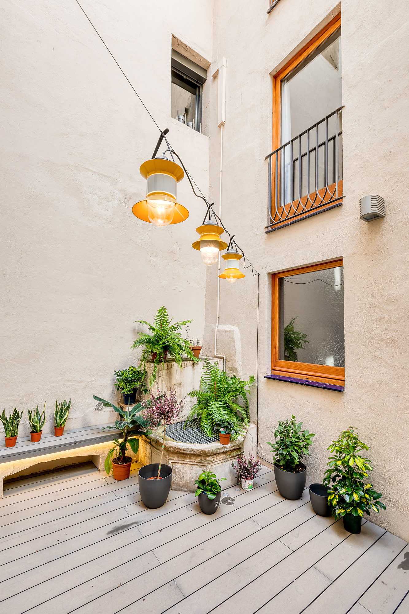 Bright and inviting private patio in a Barcelona vacation rental, featuring potted plants, wooden seating, and decorative hanging lights.