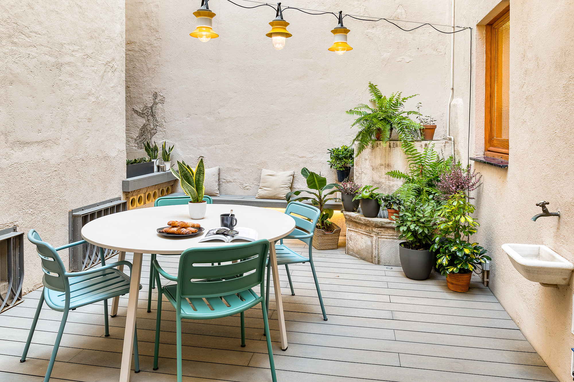 Bright and inviting private patio in a Barcelona vacation rental, featuring a round dining table surrounded by teal chairs, decorative hanging lights, and various potted plants.