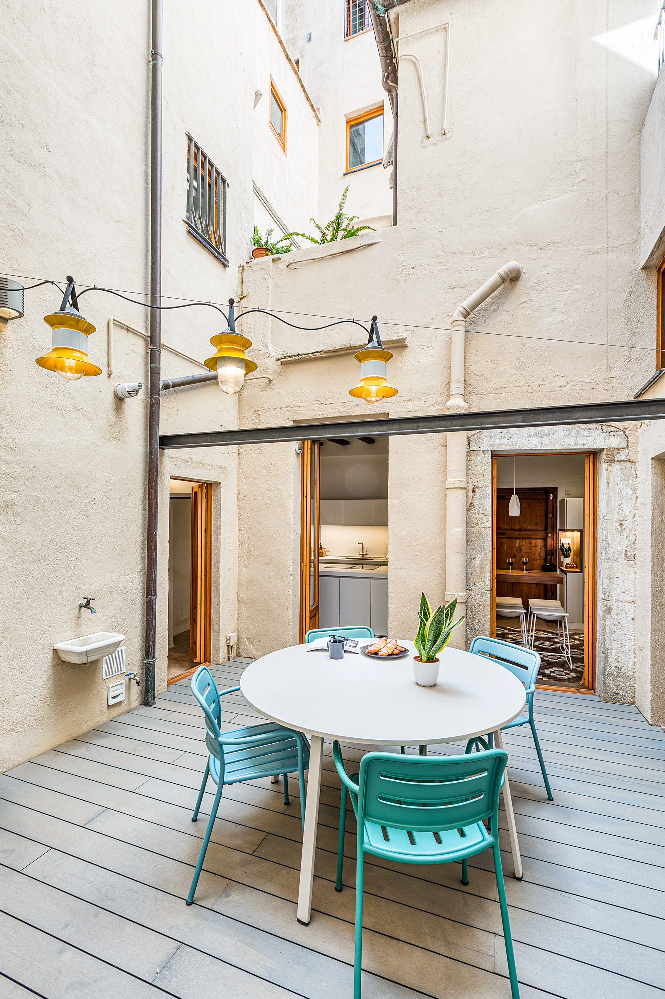 Bright and inviting private patio in a Barcelona rental, featuring a round dining table surrounded by teal chairs, with decorative hanging lights and access to the kitchen.