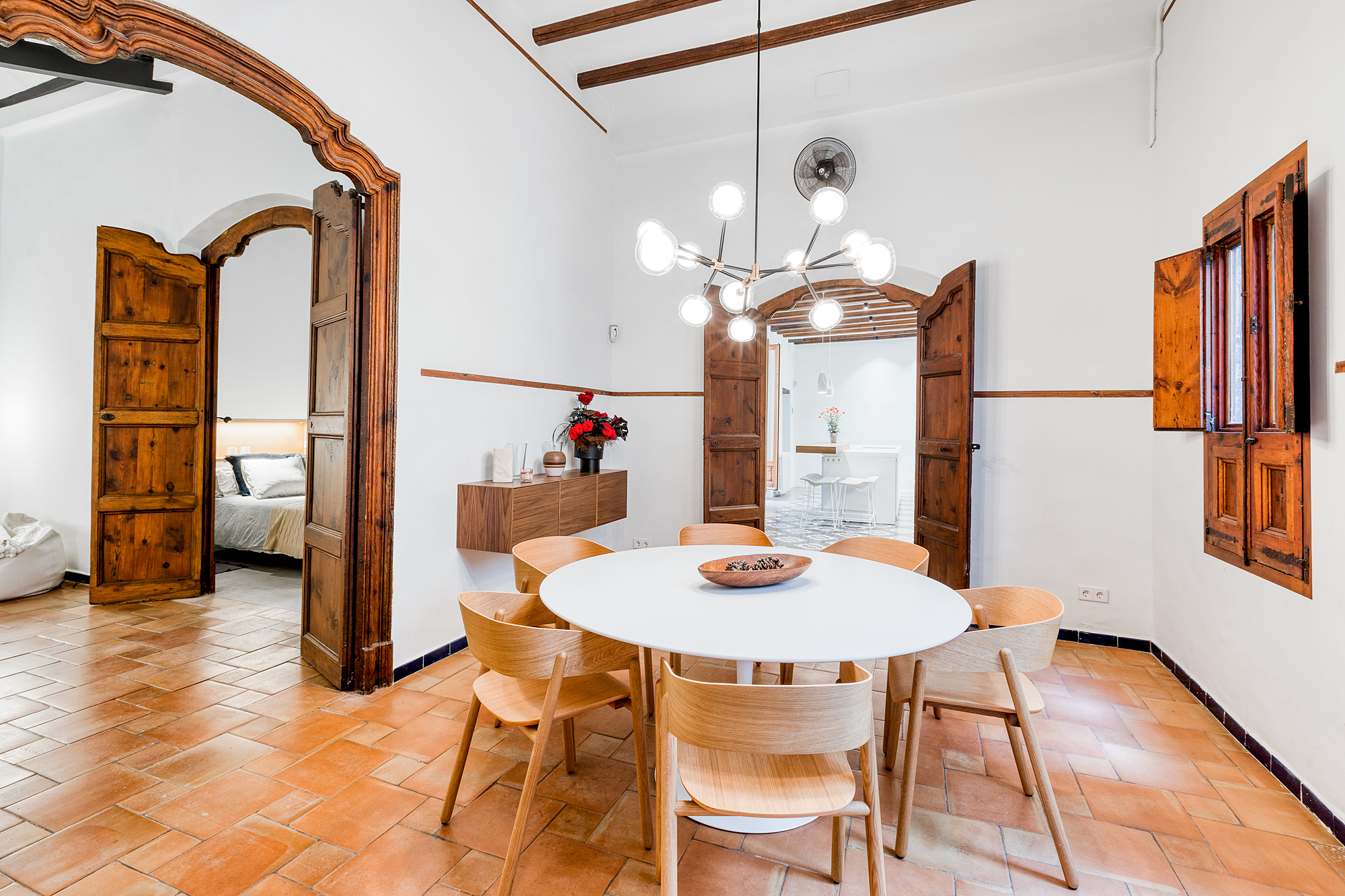 Bright dining area in a historic Barcelona home, featuring a round table with wooden chairs, decorative lighting, and terracotta flooring.