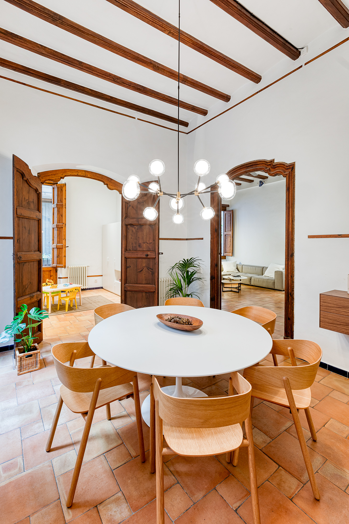Modern dining area featuring a round white table surrounded by wooden chairs, with a contemporary chandelier overhead, set in a historic Barcelona rental with terracotta flooring.