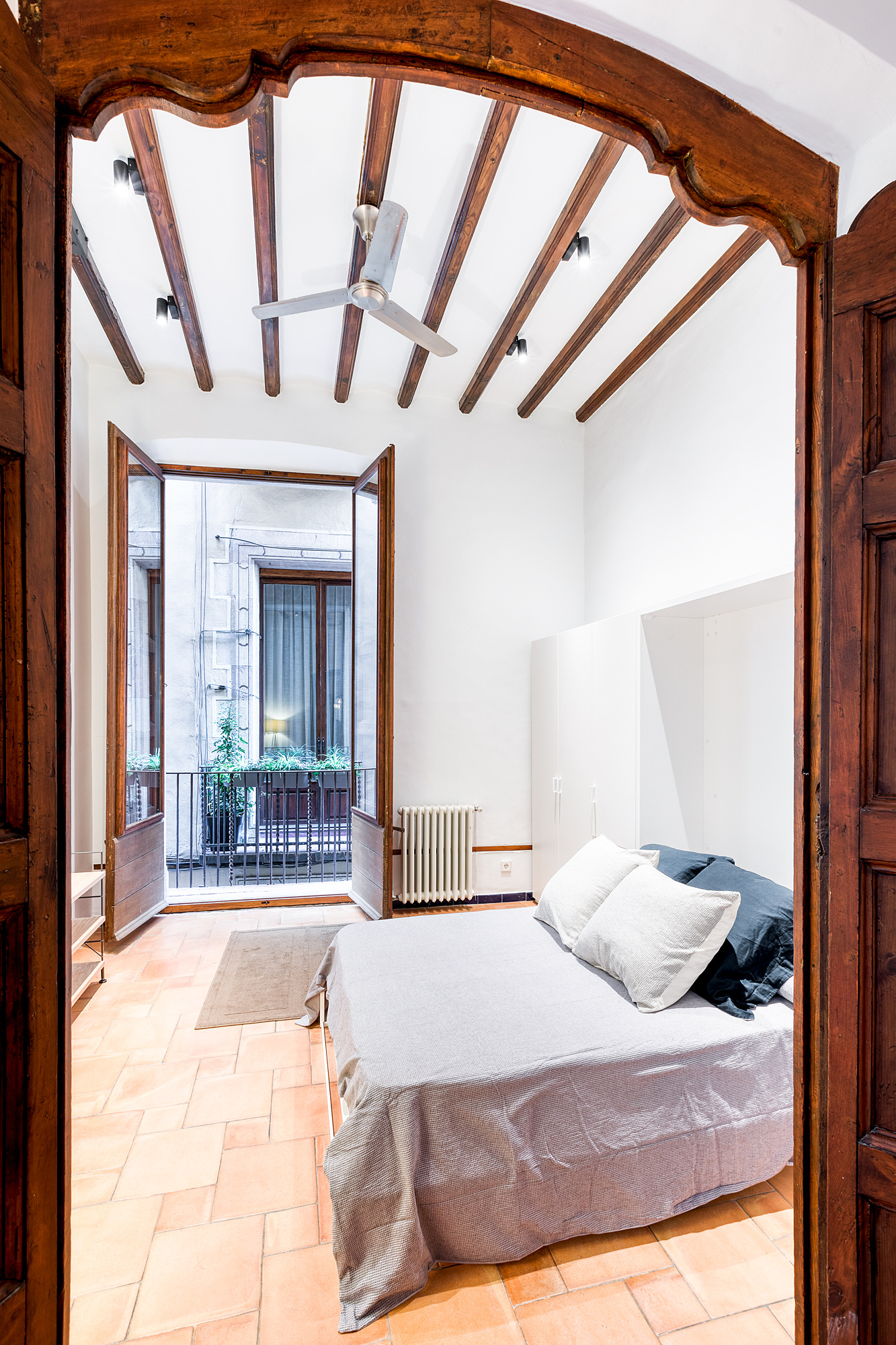 Cozy guest room in a historic Barcelona rental, featuring a queen-sized bed, wooden beams, and large doors opening to a balcony with views of greenery.
