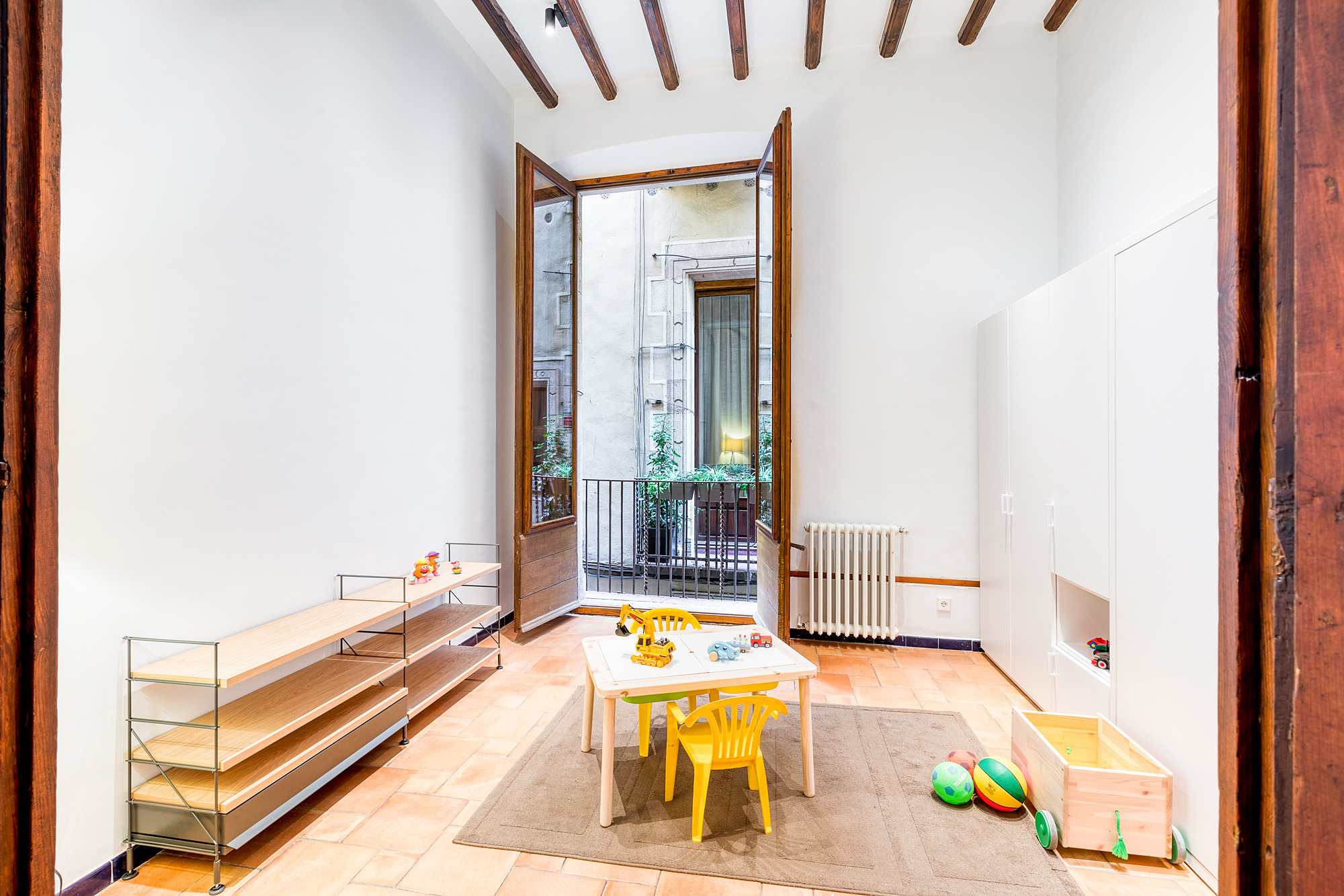 Bright playroom in a historic Barcelona vacation rental, featuring a table with toys, yellow chairs, and large windows opening to a balcony with greenery.