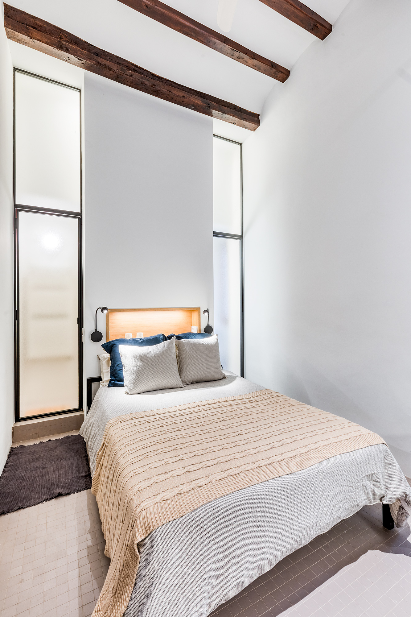 Cozy bedroom in a historic Barcelona rental featuring a queen-sized bed, decorative pillows, exposed wooden beams, and large windows for natural light.