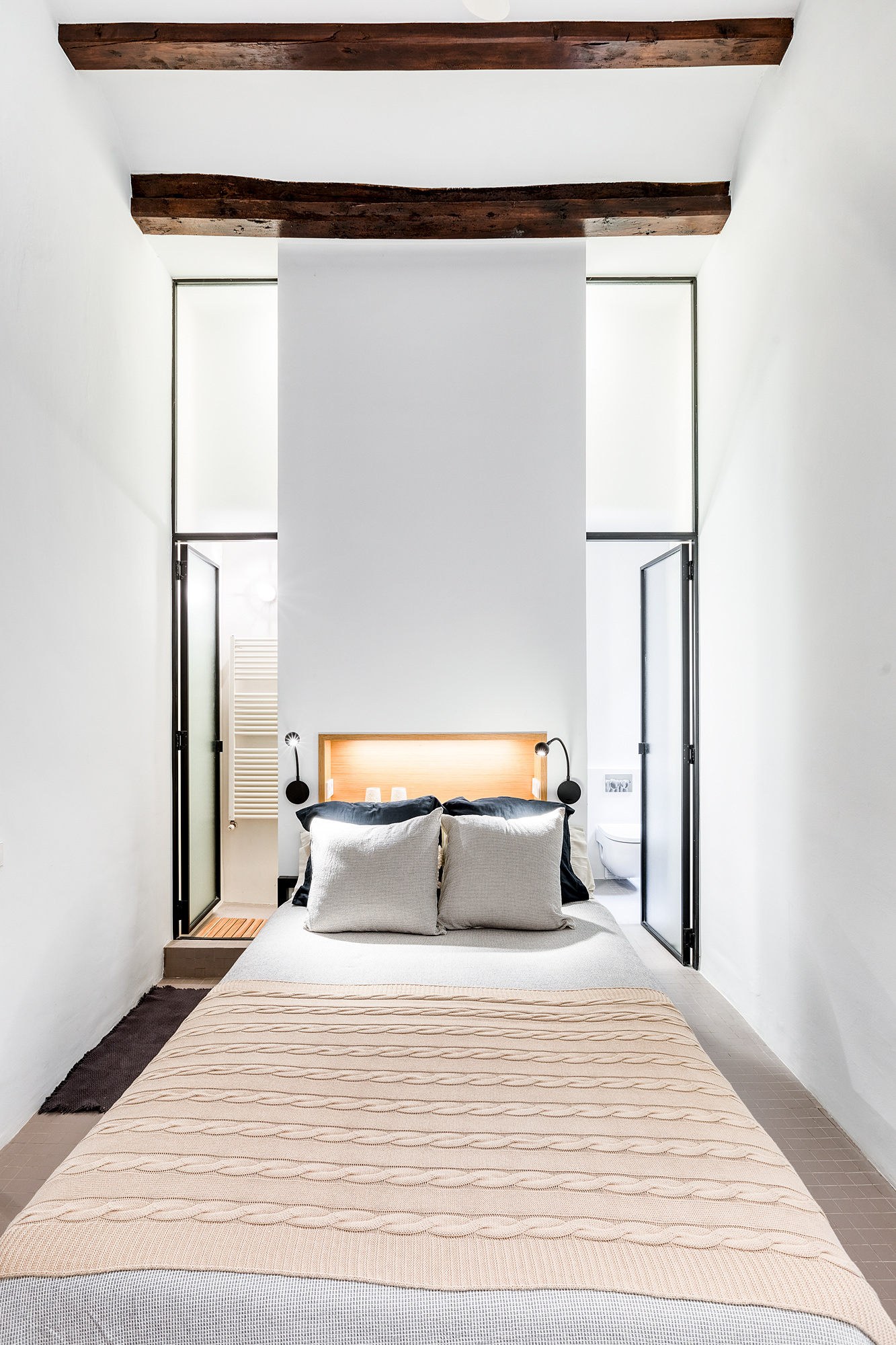 Cozy bedroom in a historic Barcelona vacation rental, featuring a queen-sized bed with decorative pillows, wooden beams overhead, and modern ensuite bathroom doors on either side.