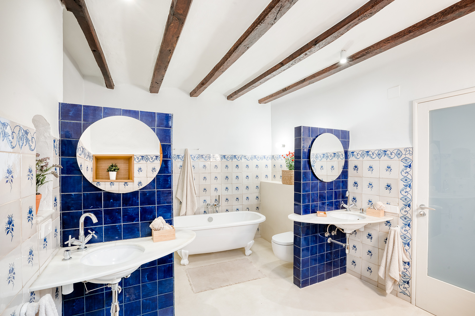 Bright bathroom featuring traditional blue and white tiles, two sinks with round mirrors, a freestanding bathtub, and wooden beams overhead.