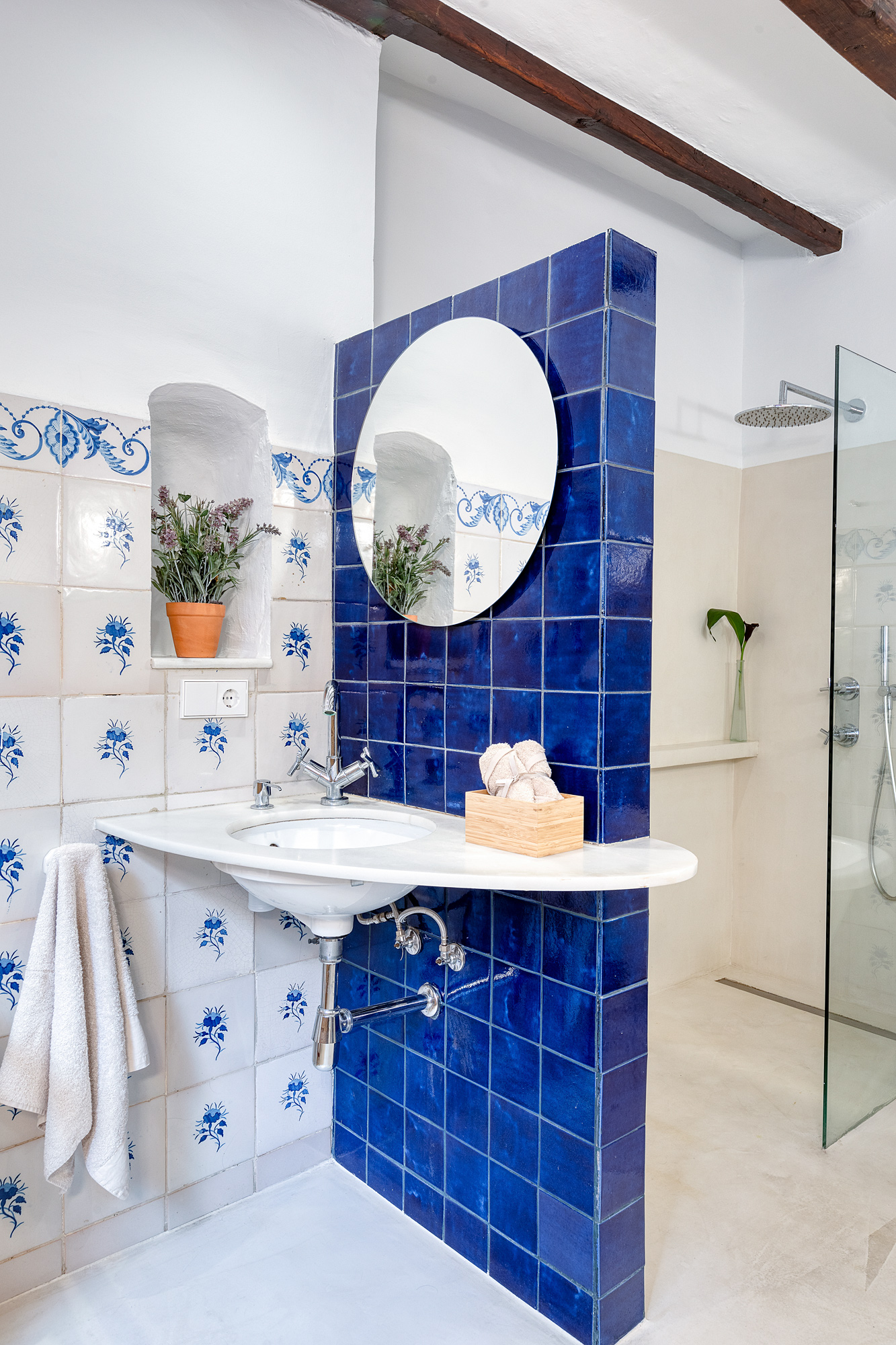 Bright bathroom showcasing blue and white tiles, with a modern sink, round mirror, and a glass shower, complemented by wooden beams.