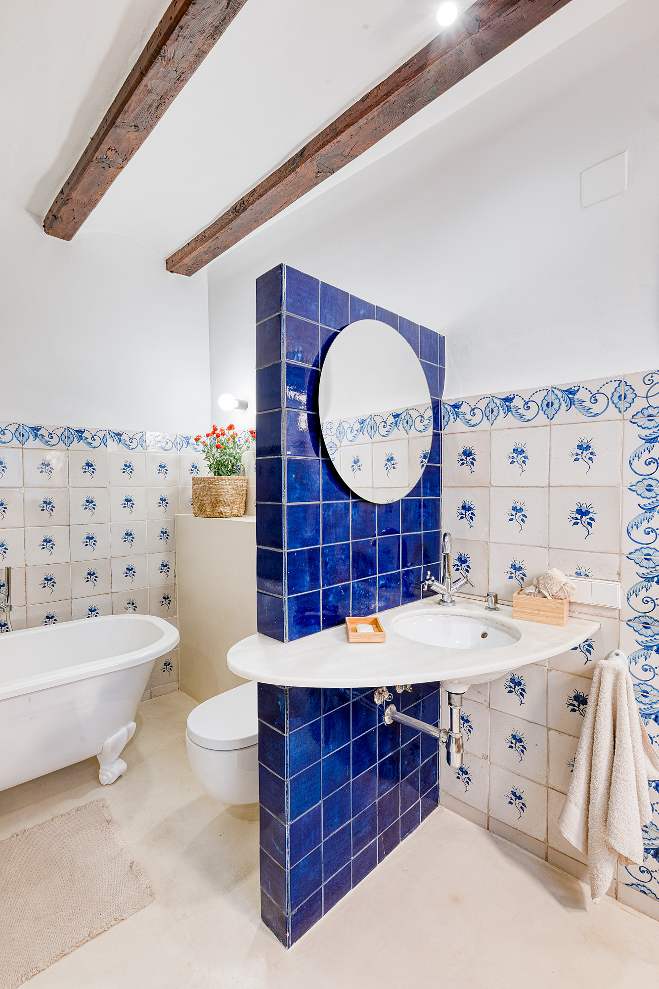 Bright bathroom featuring traditional blue and white tiles, a freestanding bathtub, a wall-mounted sink, and wooden beams overhead.