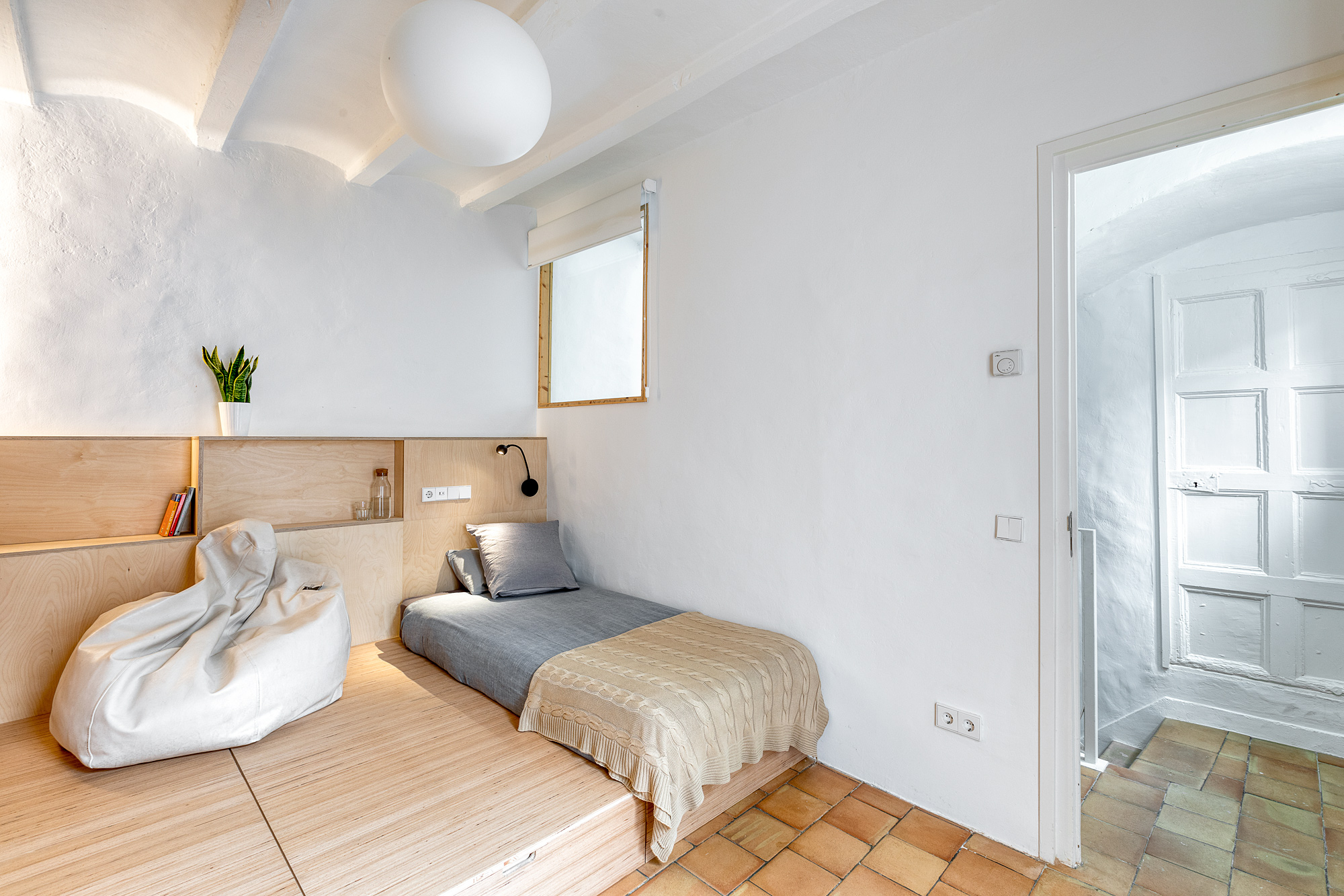 Cozy bedroom in a Barcelona vacation rental featuring a minimalist design, a bed with gray bedding, light wooden accents, and a small potted plant.