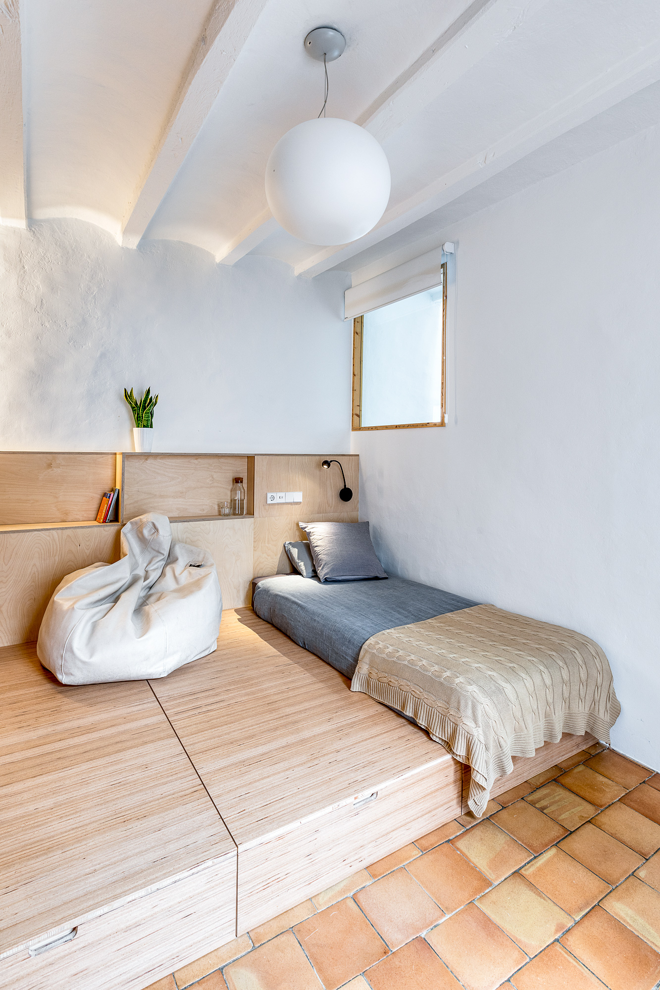 Cozy bedroom in a Barcelona vacation rental featuring a single bed with gray bedding, a bean bag chair, and minimalist wooden furnishings, complemented by a large round ceiling light and a small window.