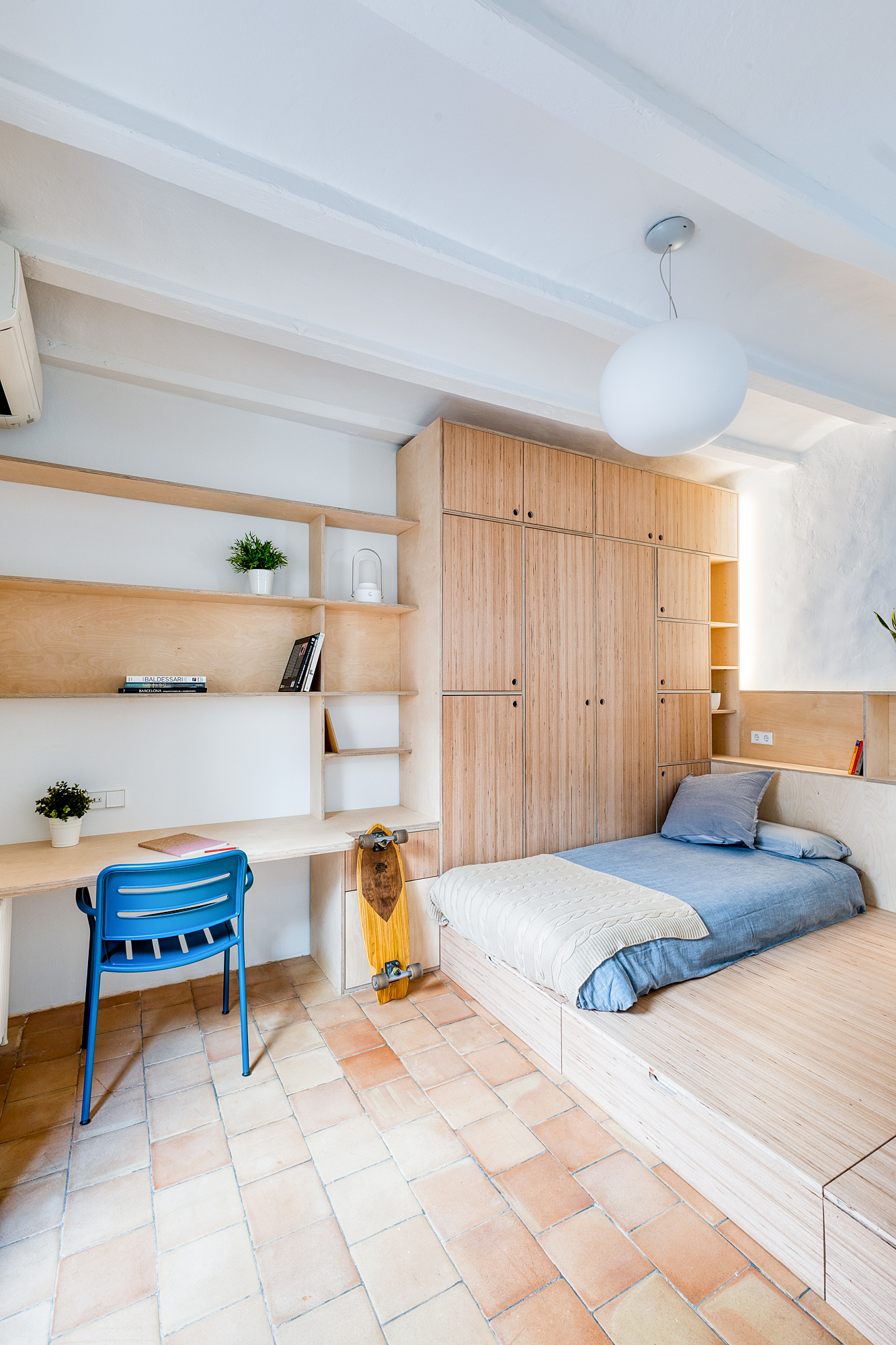 Cozy bedroom in a Barcelona vacation rental featuring a wooden wall, a queen-sized bed with blue bedding, a study desk with a blue chair, and ample natural light on terracotta flooring.