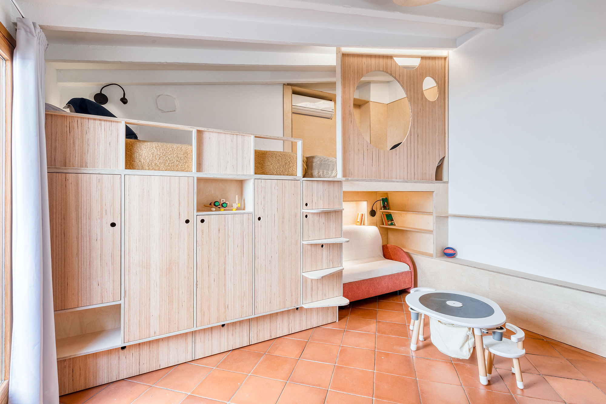 Cozy playroom in a historic Barcelona rental, featuring a wooden storage unit with shelves and hidden sleeping areas, a small round table, and terracotta flooring.