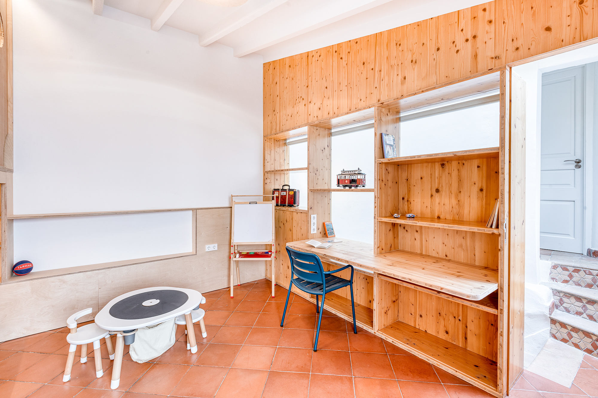 Bright playroom featuring a small table with chairs, a wooden study desk, shelves with decorative items, and terracotta flooring in a stylish Barcelona vacation rental.