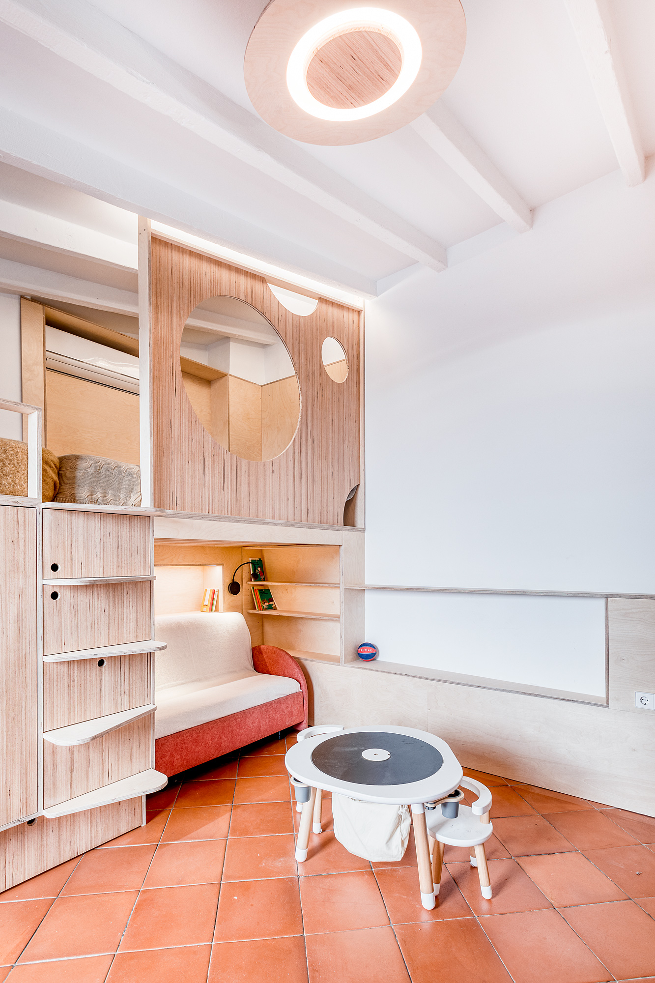 Cozy playroom in a historic Barcelona rental featuring a wooden storage unit, a red daybed, and a round children's table with chairs, complemented by a circular ceiling light.