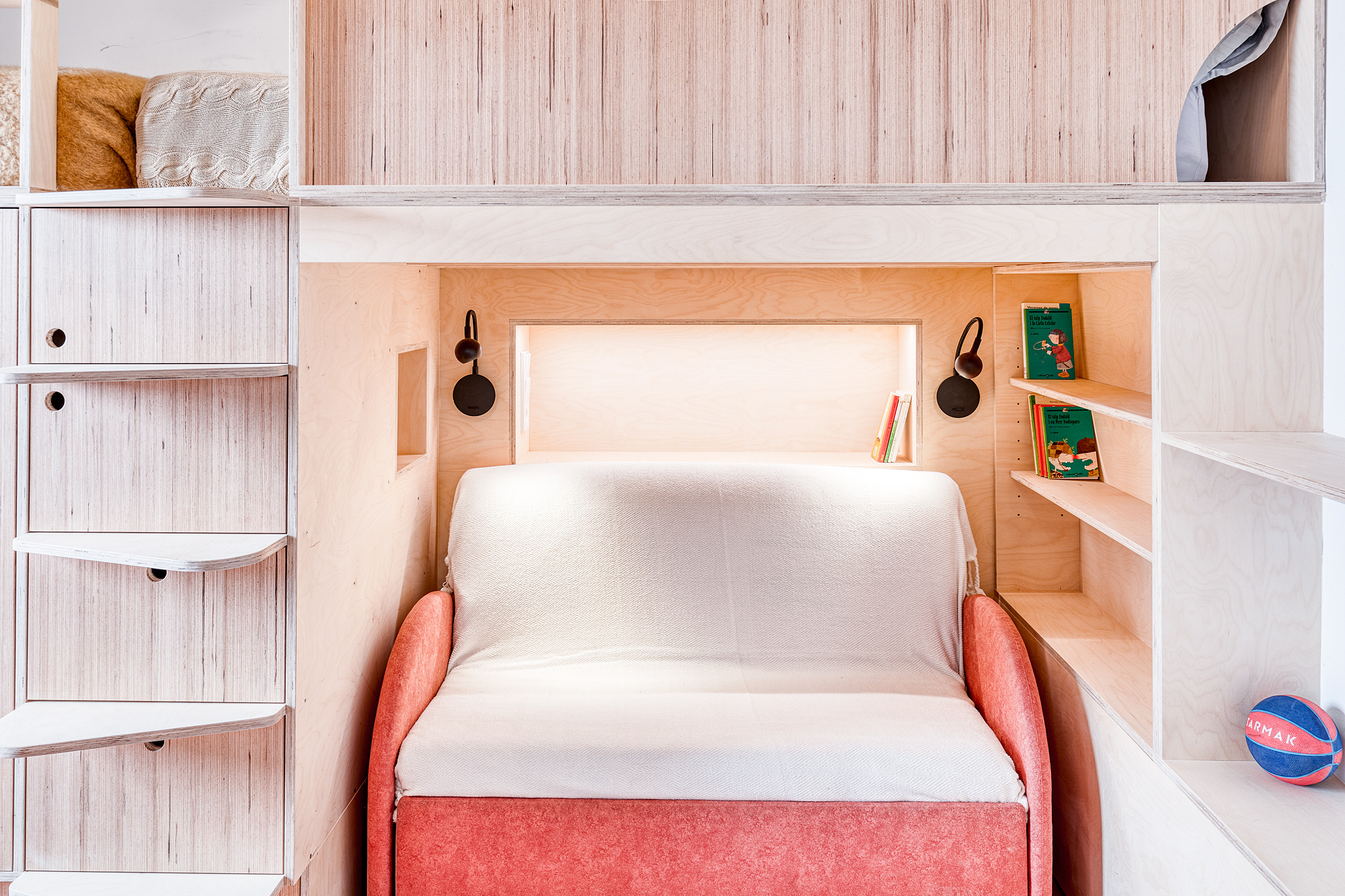 Cozy playroom in a Barcelona vacation rental featuring a convertible sofa bed with a light throw, wooden shelves with books, and soft lighting.