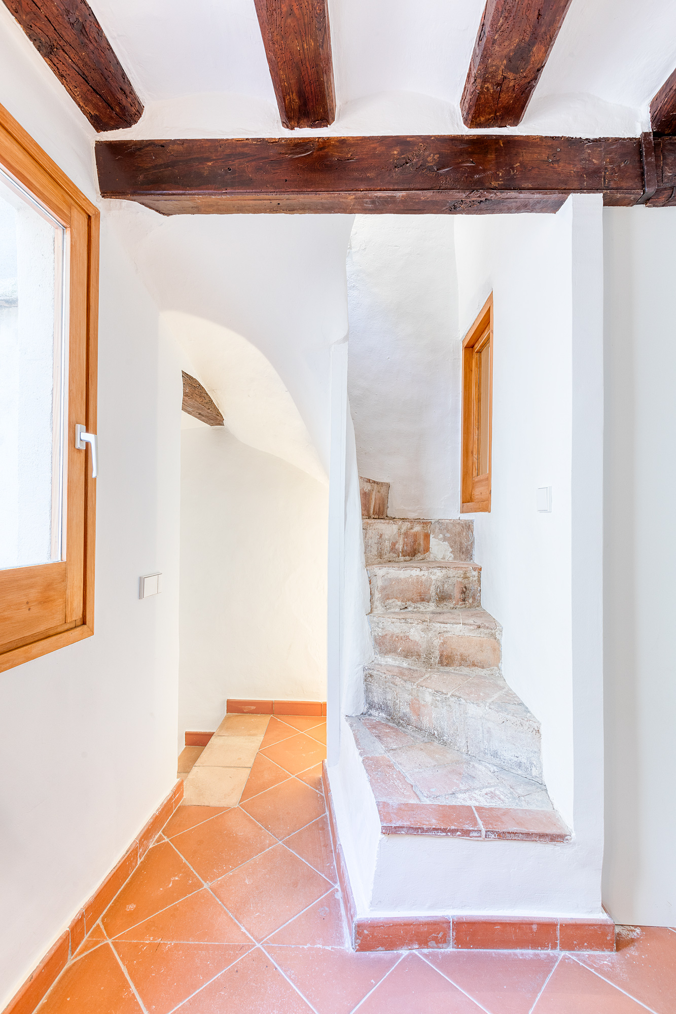 View of a historic staircase in a Barcelona vacation rental, featuring rustic terracotta steps, wooden beams overhead, and natural light from a nearby window.
