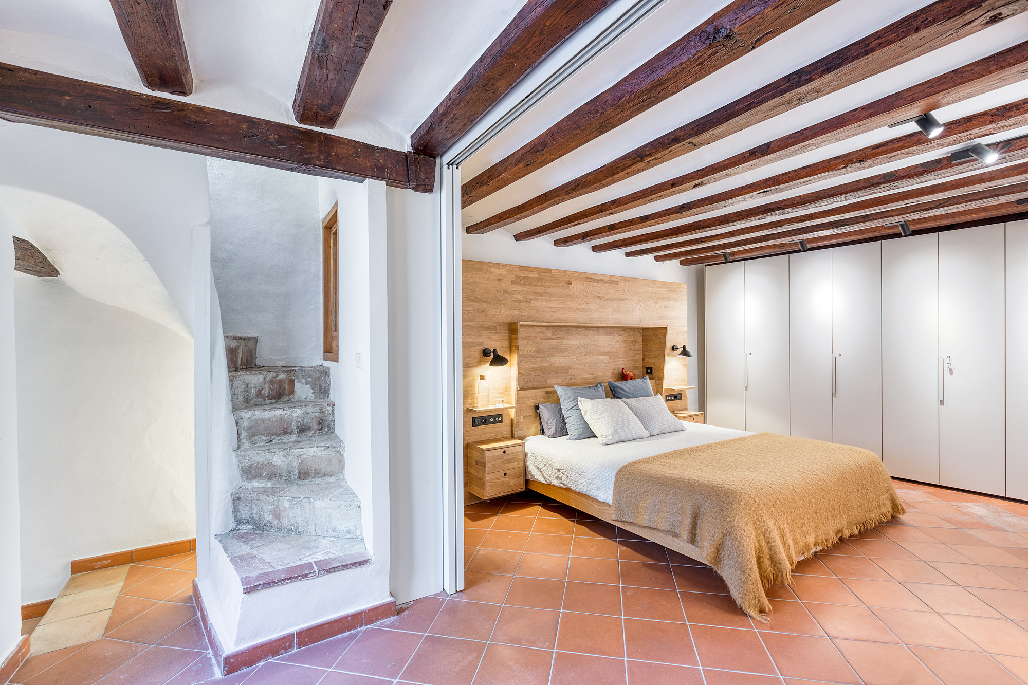 Cozy bedroom in a historic Barcelona vacation rental featuring a queen-sized bed with decorative pillows, wooden beams overhead, and a staircase leading to another level.