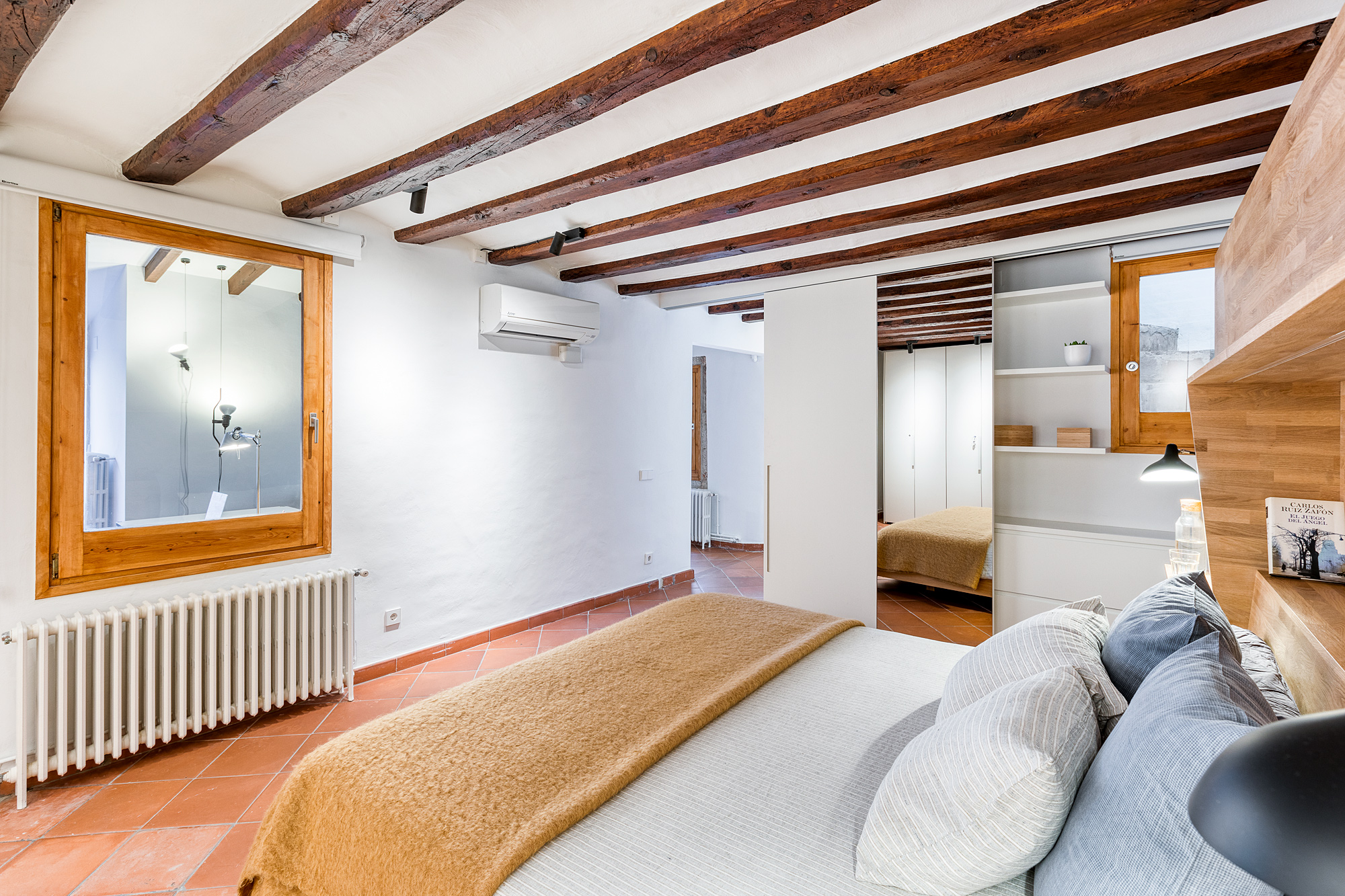 Cozy bedroom in a historic Barcelona vacation rental, featuring a queen-sized bed with decorative pillows, wooden beams overhead, and a mirror reflecting ambient light.