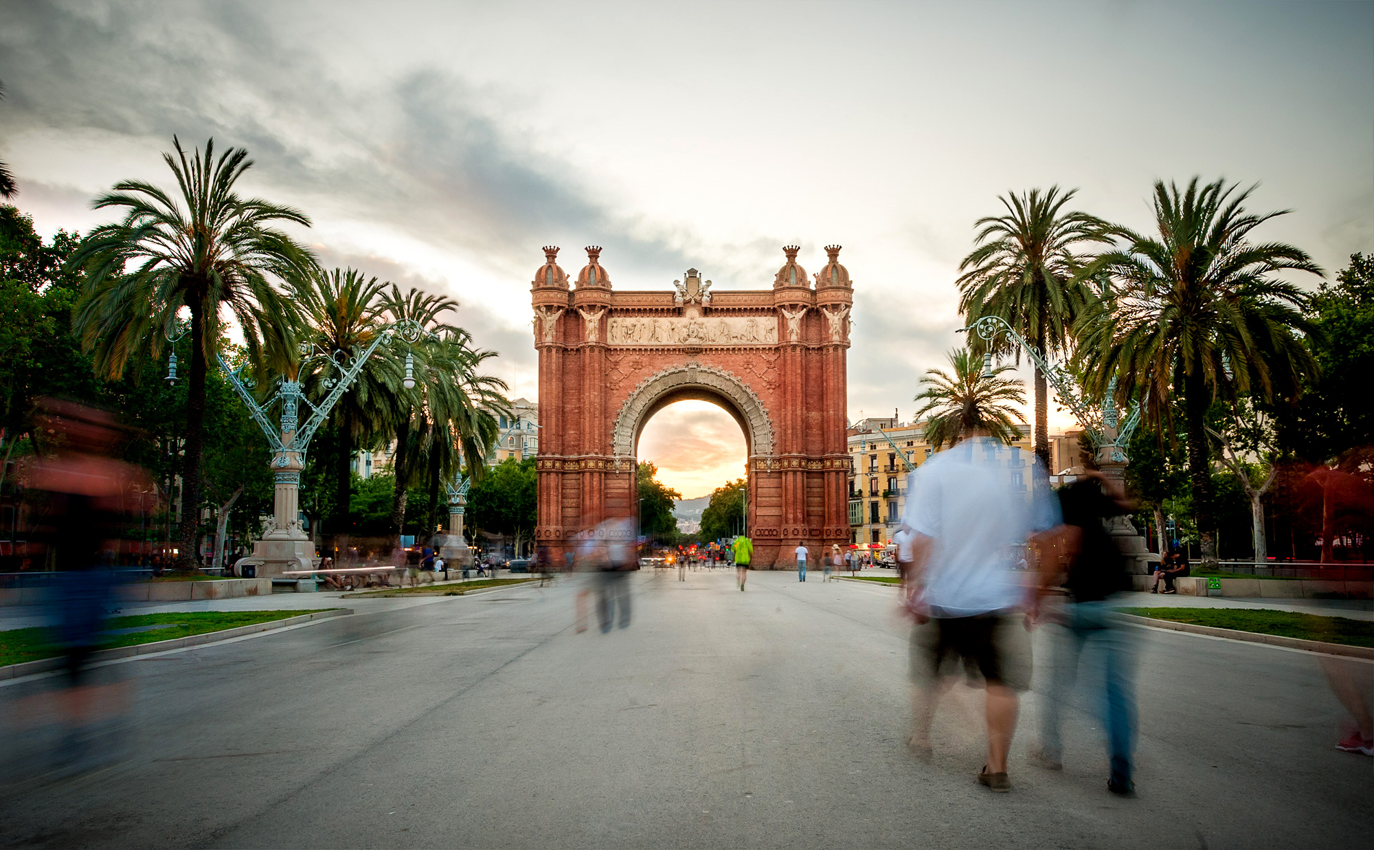 A bustling view of the Arc de Triomf in Barcelona, framed by palm trees, with people walking along a wide street during sunset.